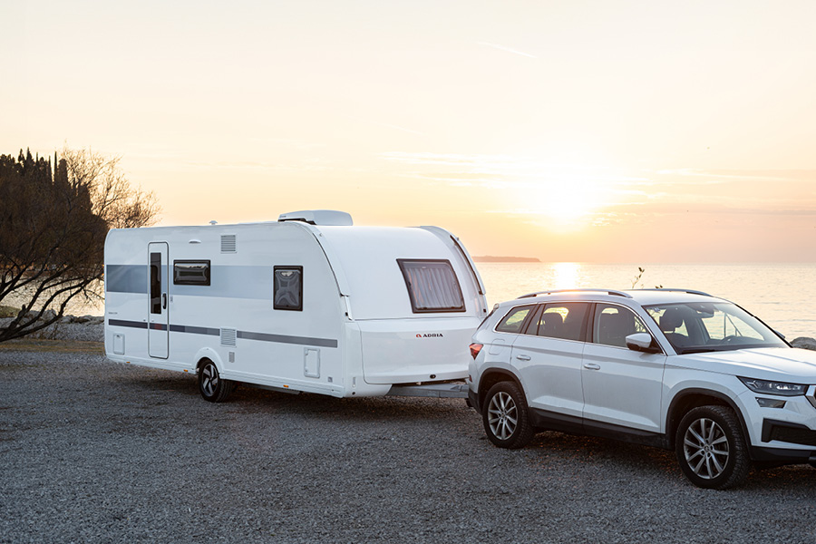 En Adria campingvogn trukket af en hvid SUV står parkeret ved en søbred i solnedgangen. Campingvognen har et stilrent design med grå og hvide striber, og der ses træer og vand i baggrunden. Det gyldne lys fra solnedgangen skaber en fredelig og idyllisk stemning.