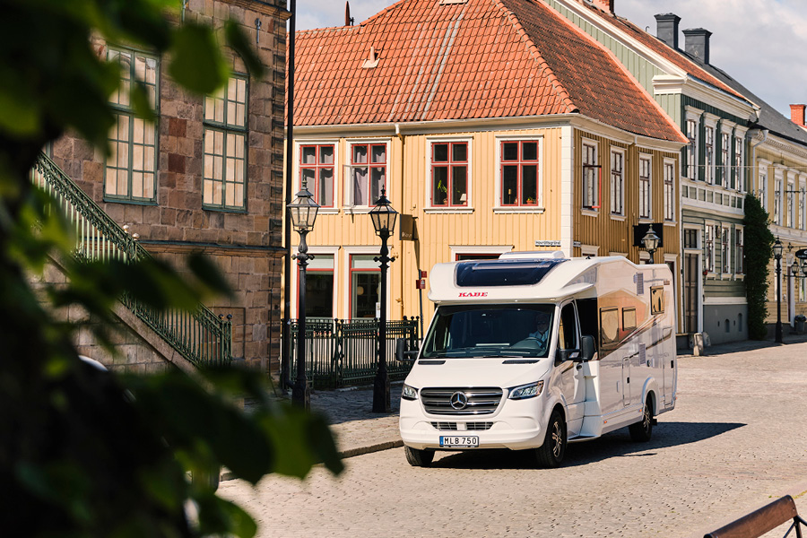 En KABE Novum 2024 autocamper parkeret på en brostensbelagt gade i en charmerende by med farverige historiske bygninger i baggrunden. Autocamperens elegante og moderne design fremhæves i det solrige vejr, hvilket viser dens alsidighed og stil.